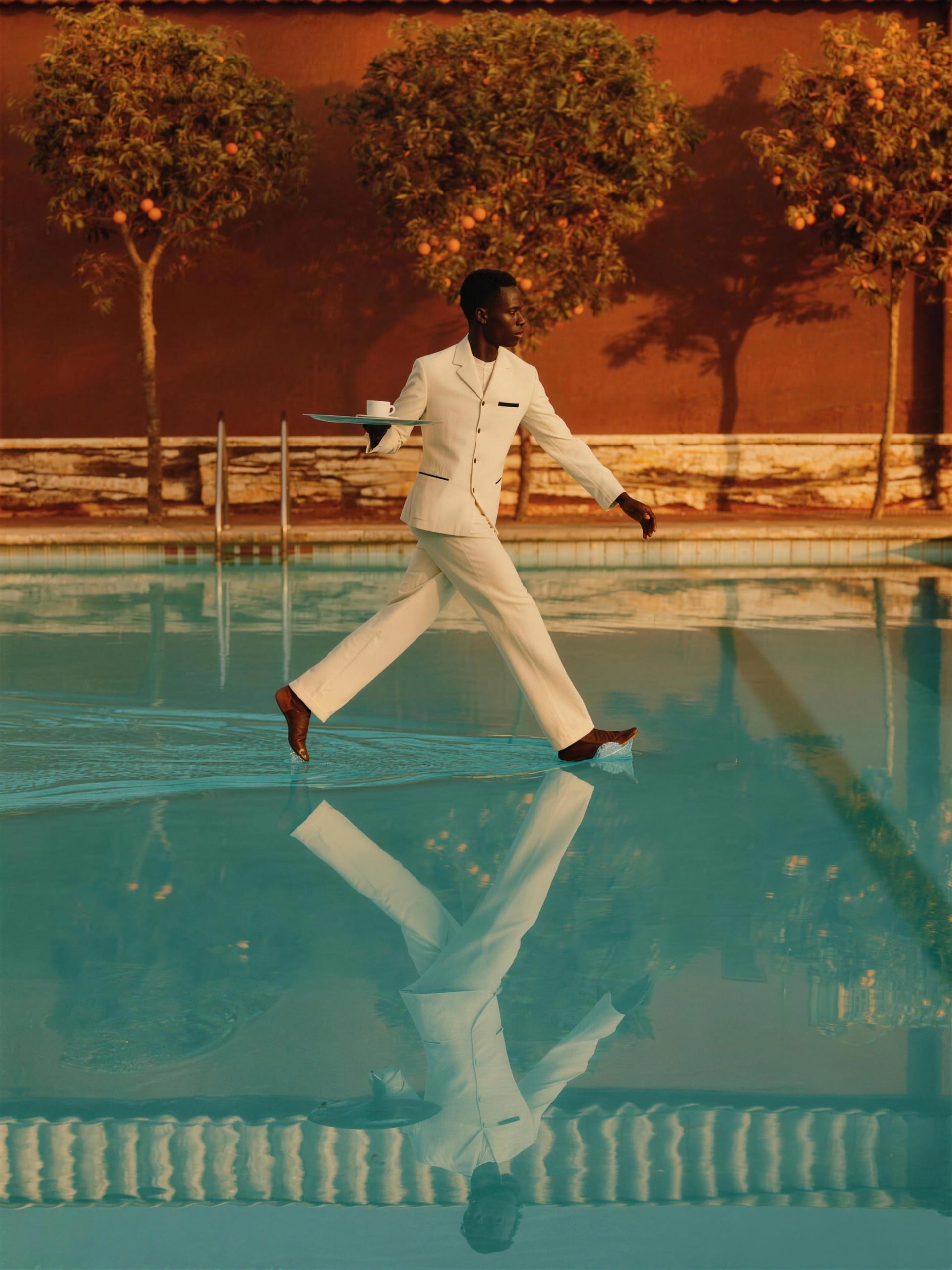 A waiter crossing a villa pool at knee depth, tray held dead level, service continuing.