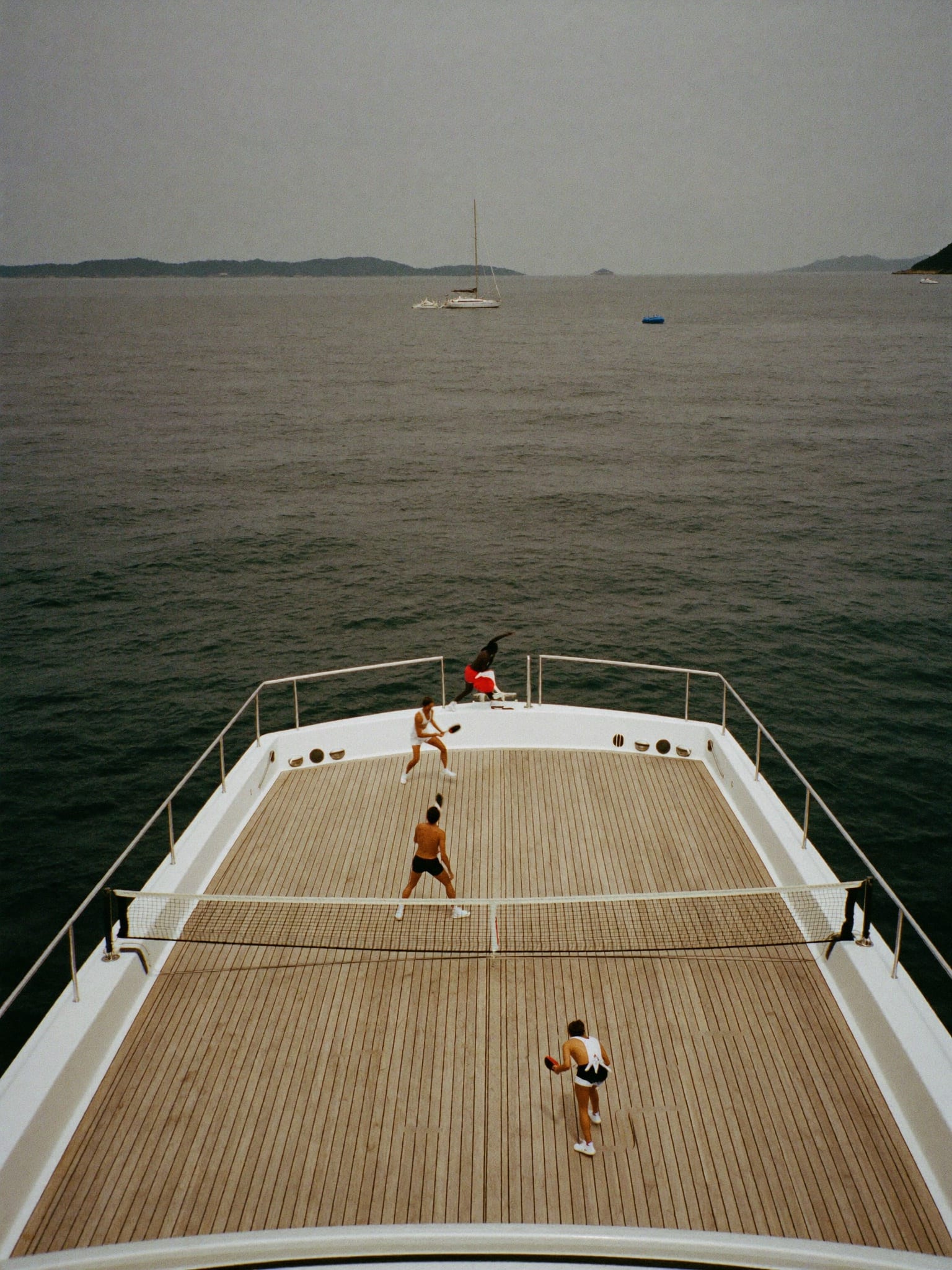 A paddle court rigged on the teak foredeck of a yacht, four players mid-rally, open sea beyond.