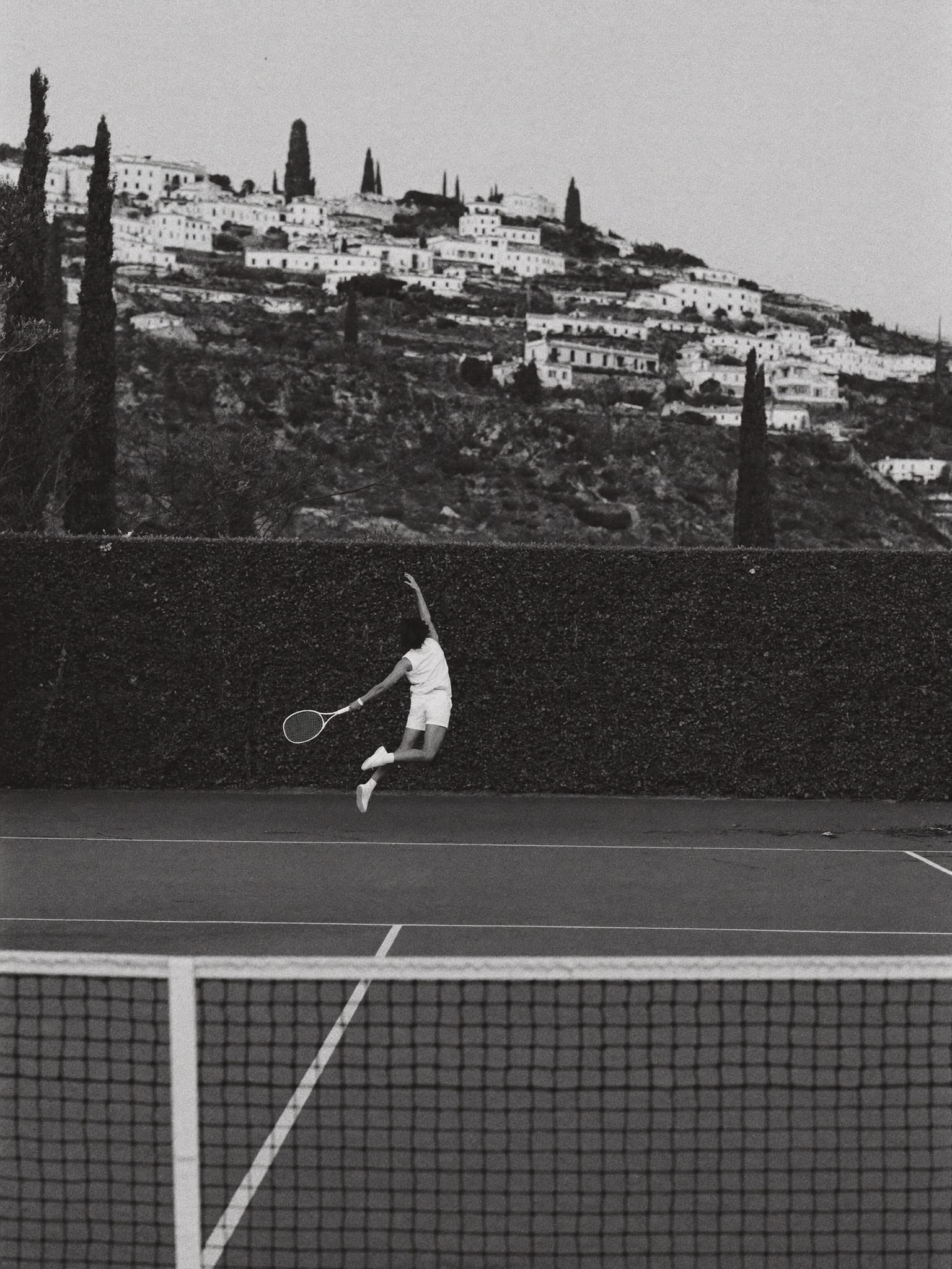A tennis player in white mid-serve on an empty court, a terraced hillside village rising behind the hedge.