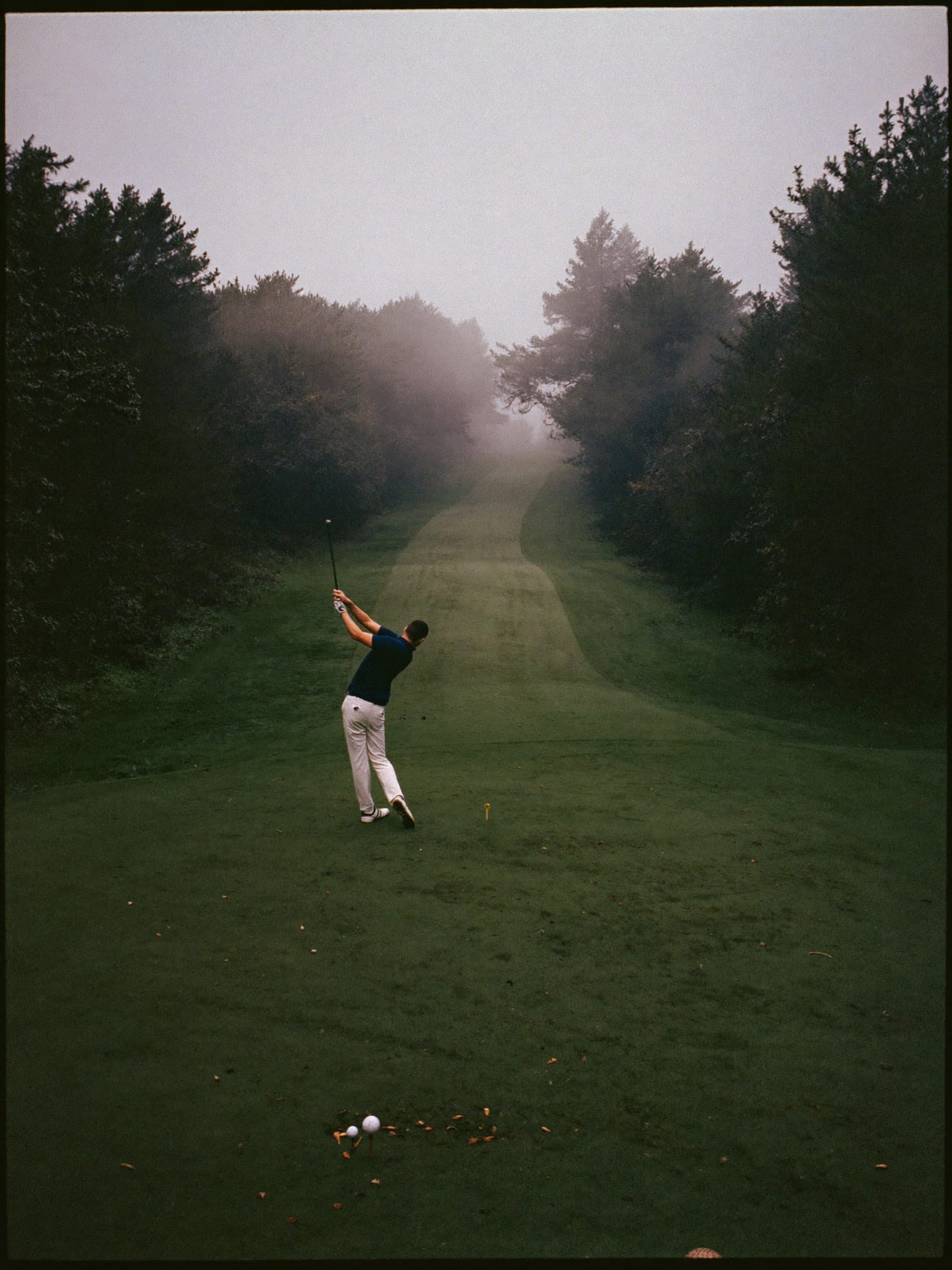 A lone golfer at the top of his swing at first light, the fairway disappearing into low morning mist.