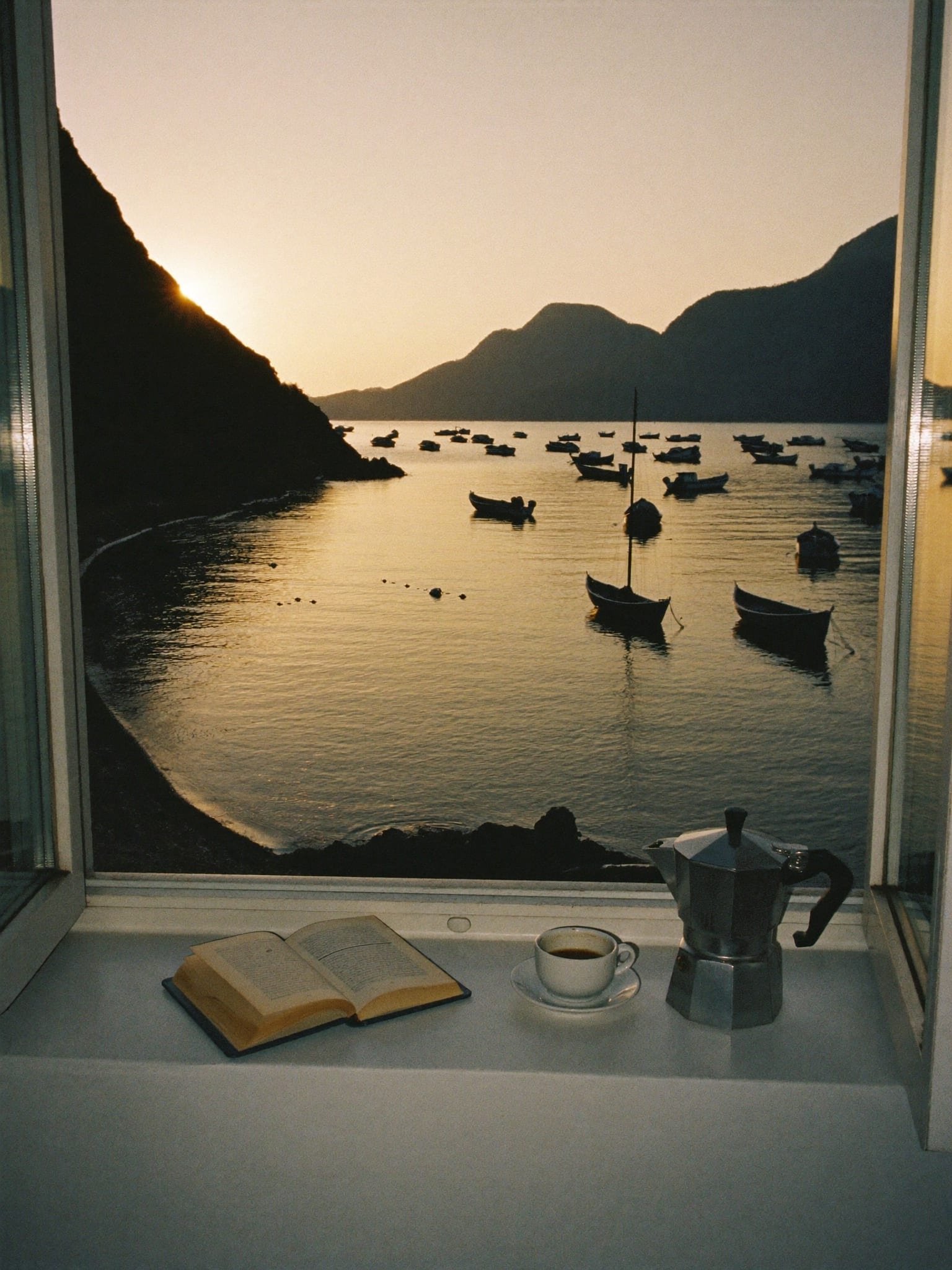 An open seaside window at golden hour, a moka pot and an open book on the sill, the bay full of moored boats.