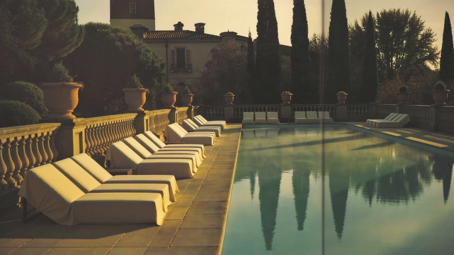 Rows of dressed sun loungers beside a long villa pool at golden hour, cypresses held still in the water.