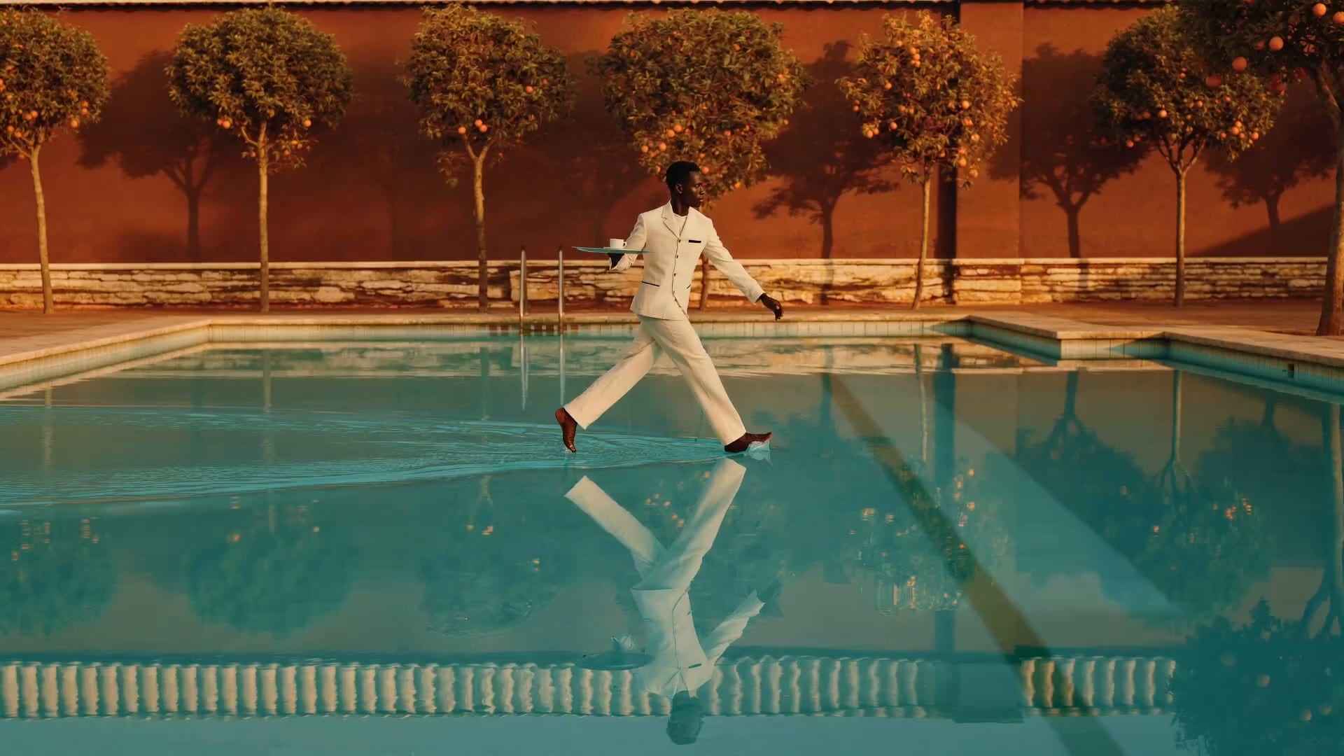 A waiter crossing the water of a villa pool, tray held level, ripples spreading from each step